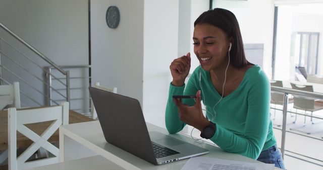 A young woman is using a smartphone while working on her laptop in a bright and modern home office. She is wearing earphones and smiling, indicating a positive conversation or productive work session. The photo conveys the concept of remote work, home office environment, and use of technology for communication. This image is suitable for illustrating concepts of remote work, modern technology, communication, and work-life balance in blogs, websites, and marketing materials.