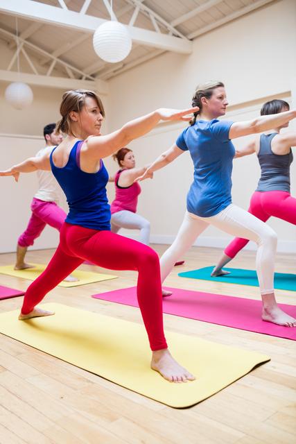 Group of individuals practicing yoga poses in a fitness studio. Participants are standing on yoga mats, and positioned in a lunge with arms extended, demonstrating focus and balance. Use this image for promoting yoga classes, fitness programs, wellness blogs, or advertisements for activewear and fitness equipment.