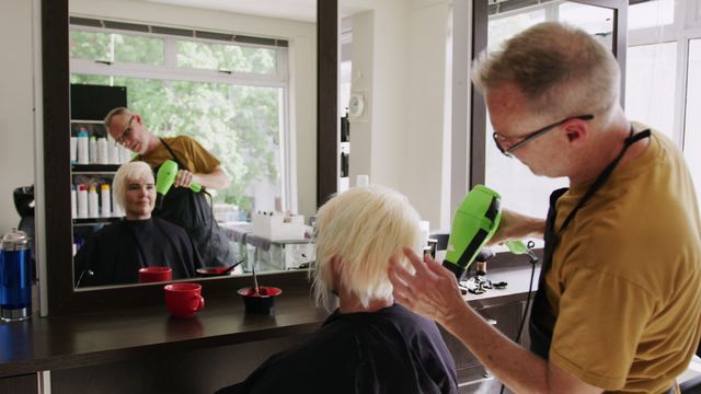 Scene captures male hairdresser expertly drying female client's blonde hair in a salon, focusing on reflection in mirror to highlight technique and teamwork. Perfect for promotions related to hair care services, salon equipment, and beauty tutorials.