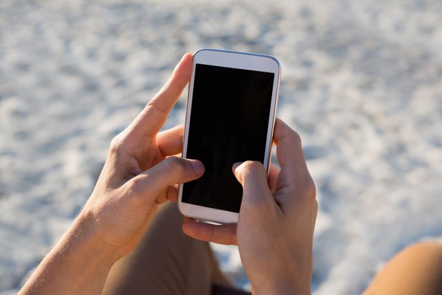 Hands holding smartphone at beach with sand in background. Ideal for themes related to summer vacations, outdoor activities, technology use in leisure time, and mobile communication. Suitable for travel blogs, tech articles, and lifestyle content.