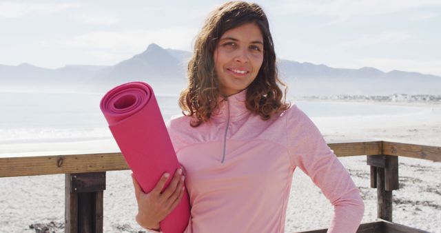 Woman holding a pink yoga mat and smiling by the beach. Ideal for promoting outdoor fitness, wellness retreats, and relaxation at coastal locations.