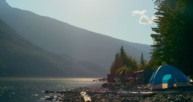 Family sitting by a tent and campfire next to a calm lake surrounded by mountains and trees. Morning sun casting golden light on the scene. Ideal for highlighting family adventure, outdoor lifestyles, camping equipment, or vacation planning.