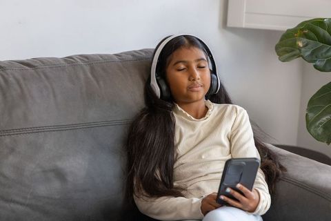 Calm asian girl relaxing on gray couch listening with headphones and holding smartphone