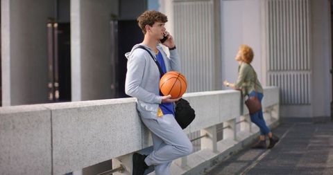 Young man leaning on concrete railing holding basketball while talking on phone in urban walkway
