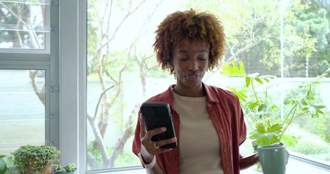 African American woman enjoying morning coffee while checking phone by sunny window