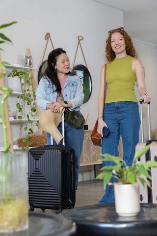 Female friends standing with rolling suitcases in stylish boutique lobby with plants