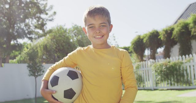 Young boy smiling while holding a soccer ball in backyard on a sunny day. Ideal for depicting themes of childhood sports, active leisure, outdoor fun, and healthy living. Useful for family, lifestyle, and sports-related content.