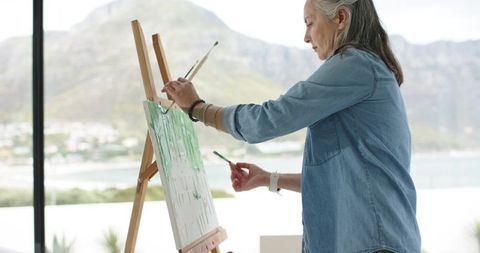 Senior woman painting on easel with coastal view, holding brushes, wearing denim shirt