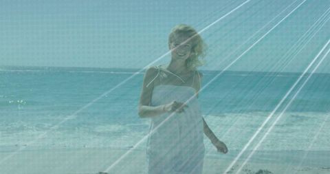 Woman walking on sunlit beach in white sundress holding smartphone, windblown hair