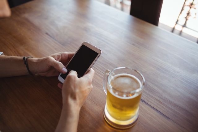 Man holding mobile phone with both hands while sitting at wooden table with glass of beer. Ideal for illustrating themes of technology use in social settings, relaxation, leisure activities, and modern lifestyle.