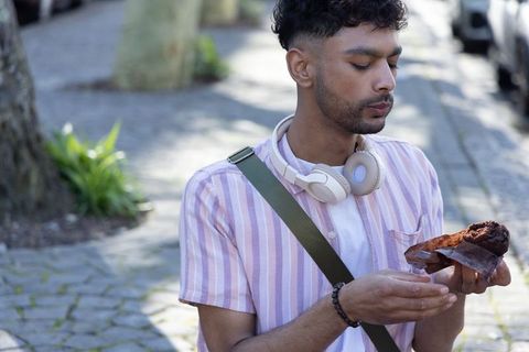 Young asian man inspecting chocolate pastry on sunlit city sidewalk wearing headphones