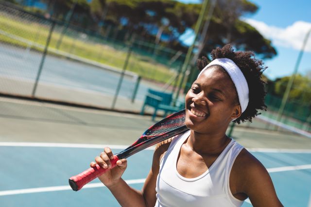 Young African American female tennis player smiling while holding a racket on her shoulder at an outdoor tennis court. Ideal for use in sports promotions, fitness campaigns, and advertisements for athletic gear or tennis equipment.