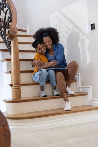 African american mother and son sharing tablet on curved wooden stairs, smiling, hugging