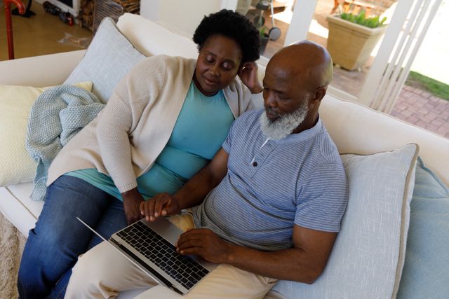 African American senior couple sitting on a couch, engaging with a laptop. The man is typing while the woman watches attentively. Ideal for use in articles or advertisements about technology use among seniors, retirement lifestyle, online communication, or home life.