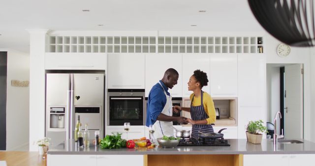 Depicts a couple happily cooking together in a contemporary kitchen, emphasizing joy and togetherness. Perfect for use in articles about relationships, healthy lifestyles, culinary blogs, cooking classes, kitchen interior designs, or digital marketing related to kitchen appliances and cookware.