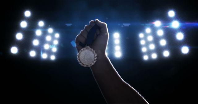 Depicts close-up of a hand raising a silver medal with bright stadium lights in the background. Ideal for use in articles or advertisements about sports achievements, competitions, winning, motivation, and celebrating success. Suitable for promoting sporting events, athlete profiles, and award ceremonies.