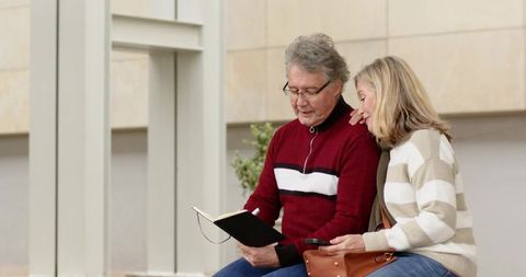 Senior couple reviewing planner on outdoor bench with smartphone and leather handbag