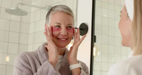 Mature woman and daughter applying pink under-eye gel patches at vanity mirror, smiling