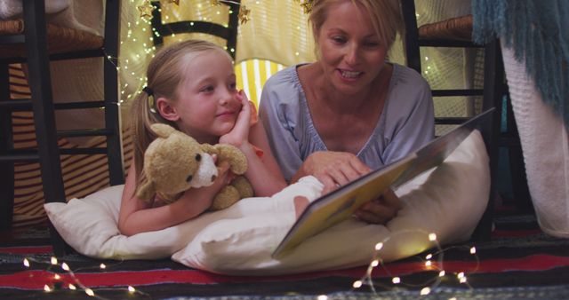 Mother engaging in bedtime reading session with young daughter in warm, homemade fort illuminated by fairy lights. Girl holding teddy bear while listening intently. Ideal for use in family-oriented publications, parenting blogs, or articles about childhood, bonding, and parenting.