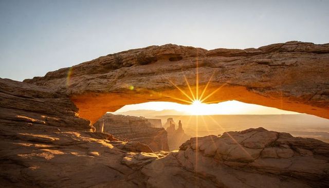 Starburst sun framing through natural sandstone arch over vast desert canyon at sunrise