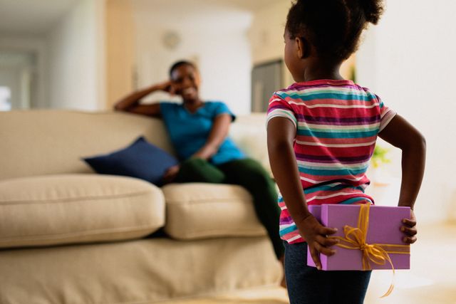 Little girl holding a gift behind her back, preparing to surprise her mother who is sitting on a couch. Perfect for themes related to family bonding, celebrations, parenting, and special moments at home.