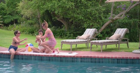 Poolside friends passing drinks and playing with beach ball on sunny backyard deck