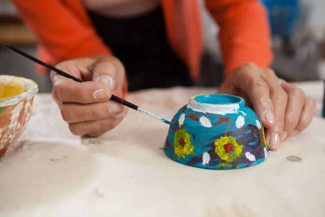 Mid section view of a woman painting a colorful ceramic bowl in an arts and crafts class. The focus is on her hands and the paintbrush as she adds intricate details to the pottery piece. This image can be used for content related to creative hobbies, art workshops, and pottery classes.