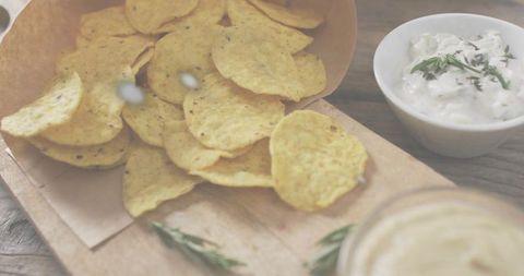 Spilling corn tortilla chips from paper cone on rustic wood board with creamy herb dip