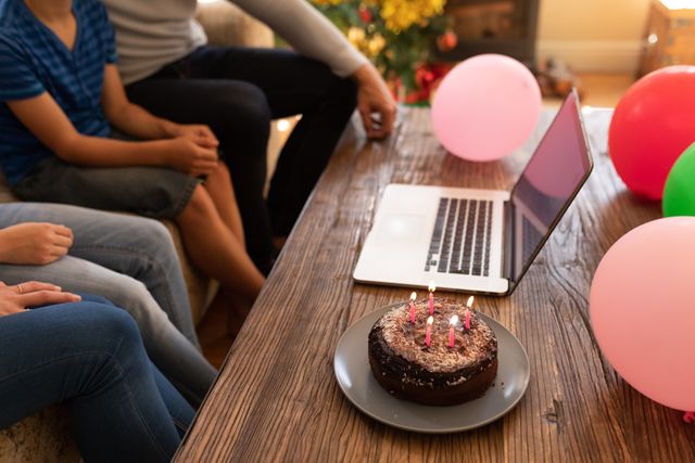 Family sitting on couch in living room, celebrating a birthday virtually with a laptop. Birthday cake with five candles and balloons on table. Perfect for themes of remote celebrations, family bonding, and virtual gatherings.
