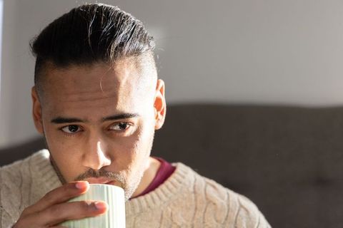 Thoughtful young man sipping coffee on sofa wearing knit sweater, cozy morning portrait, warm light