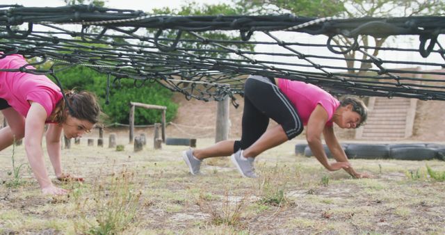 Determined caucasian female friends in pink t shirts crawling under nets on bootcamp training course. Female fitness, friendship, challenge and healthy lifestyle.