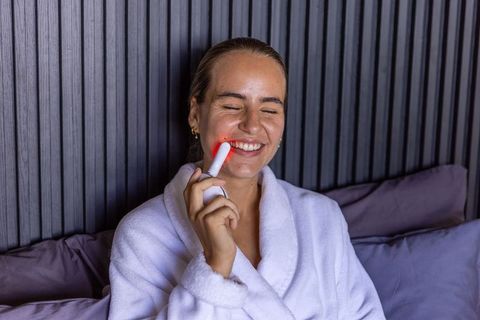 Woman using red light facial device in white bathrobe enjoying at-home skincare ritual and relaxatio