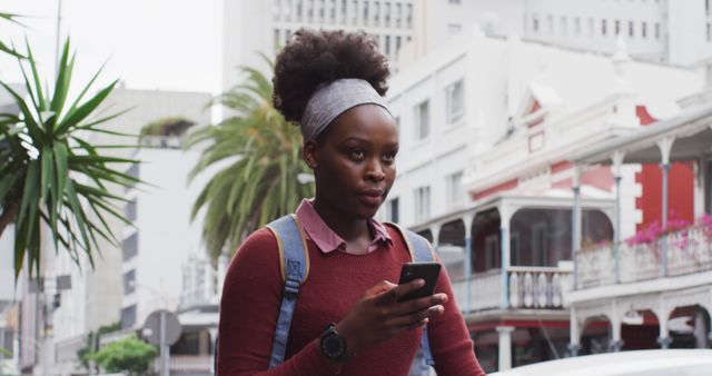 Young African American woman in casual wear stands on a city street holding smartphone, looking focused. She has a backpack, suggesting she is either a tourist or commuting. Palm trees and historic buildings provide an urban backdrop. This image is ideal for content related to technology, urban life, travel, solo adventure, and modern living.