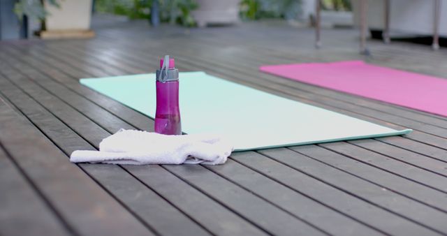 Yoga mat with a water bottle and a towel on a wooden deck. Great for using in fitness, workout, yoga class marketing materials or promotions of outdoor exercise activities.