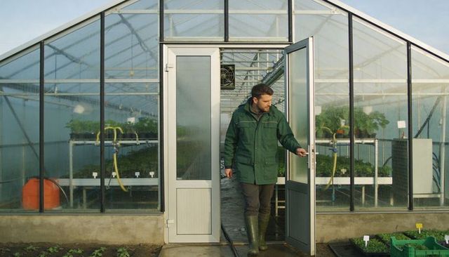 Walking greenhouse grower exiting glass door wearing green coat and rubber boots
