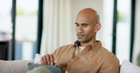 Bald man relaxing on couch using tablet for remote work, browsing, or streaming at home
