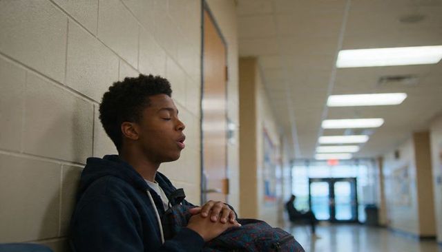 Teen student in hoodie leaning against school wall holding backpack in quiet hallway