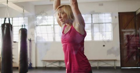Woman stretching overhead in gym with punching bags and world map overlay for fitness