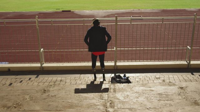 Athlete with prosthetic legs stands looking at a race track, wearing headphones. Person exhibits a motivated and determined attitude, ready for training or competition. Can be used for topics related to sports, fitness, motivation, overcoming challenges, inclusivity, and disability in athletics.