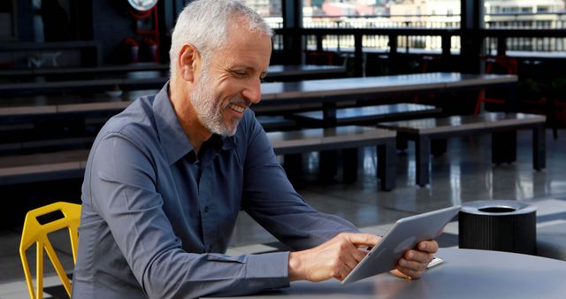 Caucasian businessman reviews digital tablet at an outdoor cafe. He displays a content smile, engaged in his work on a sunny day.
