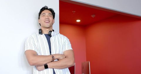 Confident asian man leaning against wall wearing headphones and smartwatch in workspace