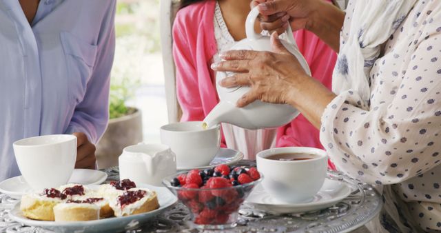 A diverse group enjoys an afternoon tea together, with copy space. Delicate hands pour tea into cups, complementing the spread of scones and fresh berries on the table.