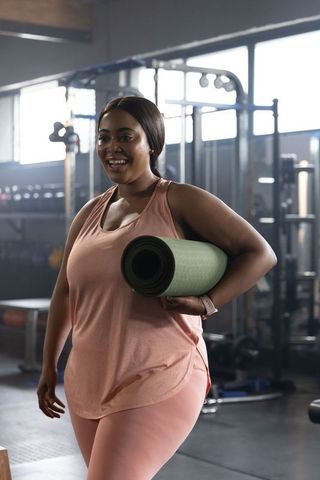 African american woman carrying green yoga mat smiling while walking through gym wearing pink gear
