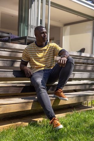 African american man sitting on outdoor deck bench wearing striped tee and sneakers