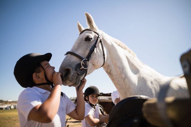 Boy kissing white horse at ranch on sunny day. Children wearing helmets, engaging in equestrian activities. Ideal for themes of animal bonding, outdoor activities, childhood experiences, and rural life. Perfect for promoting equestrian centers, summer camps, and pet care.