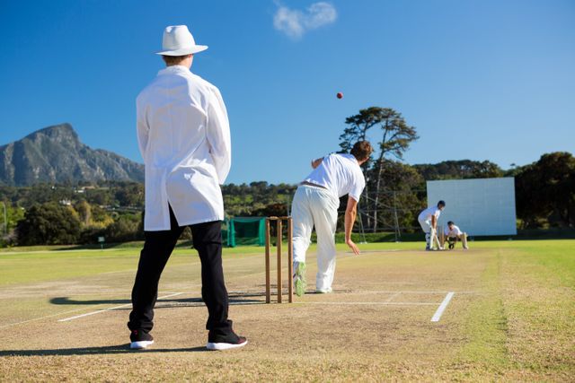 Cricket match taking place on a sunny day with an umpire overseeing the game and a bowler delivering the ball. Ideal for use in sports-related content, articles about cricket, outdoor activities, team sports, and athletic competitions.