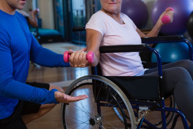This image depicts a physiotherapist assisting a senior woman in a wheelchair with a dumbbell exercise. It is ideal for use in articles or advertisements related to physical therapy, rehabilitation, elderly care, and fitness programs for seniors. The supportive interaction highlights the importance of professional guidance in maintaining health and wellness for elderly individuals.