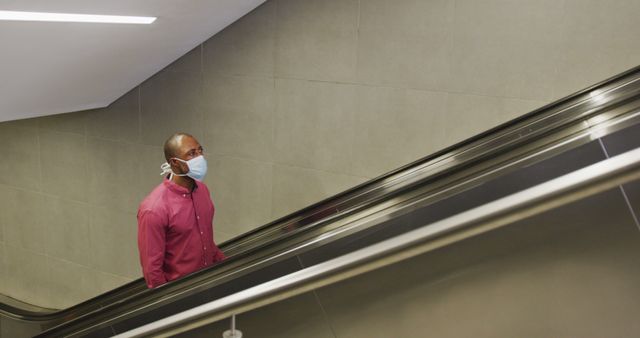 Man rides escalator in public space, wearing a face mask. Ideal for illustrating public health safety measures during the COVID-19 pandemic, social distancing protocols, and urban lifestyle scenes. Could be used in health and safety campaigns, informational content on protective measures, or related to public transport.