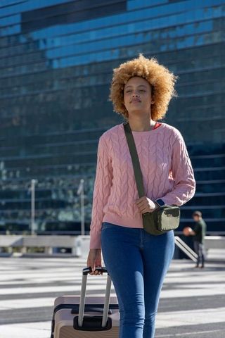 Young woman walking with suitcase and crossbody bag through glass plaza, urban travel