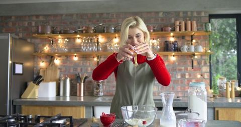 Home baker mixing ingredients cracking egg into bowl in cozy rustic kitchen with lights
